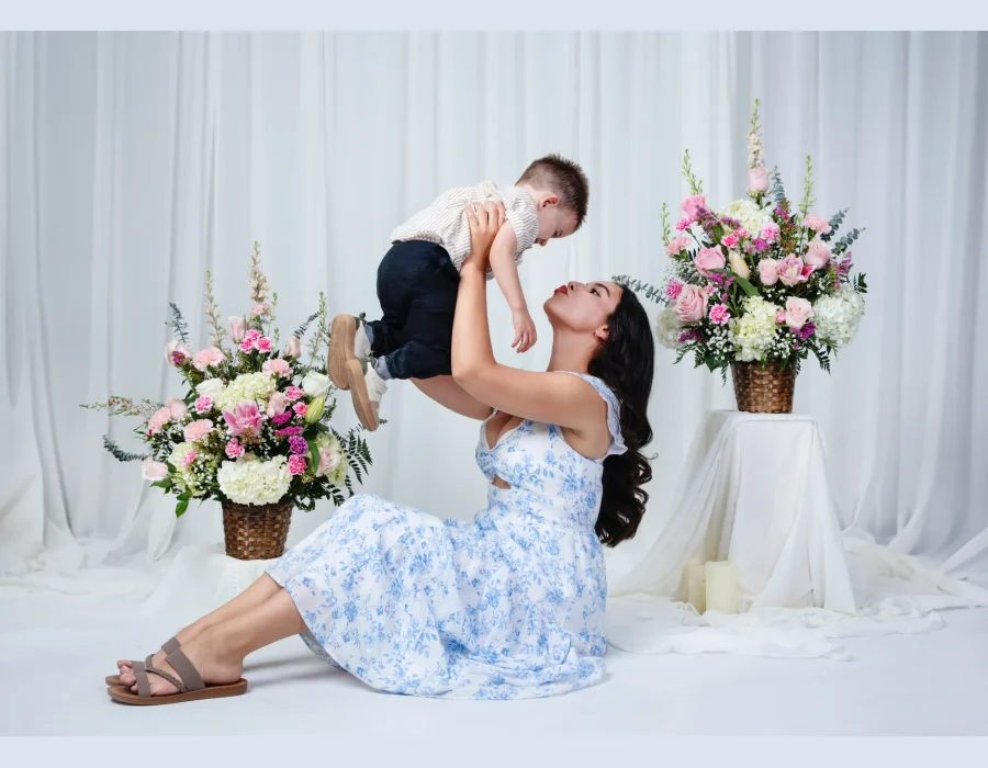 Playful portrait of a mother in a blue floral sundress sitting on the floor and lifting her toddler son overhead, surrounded by pink flower arrangements.