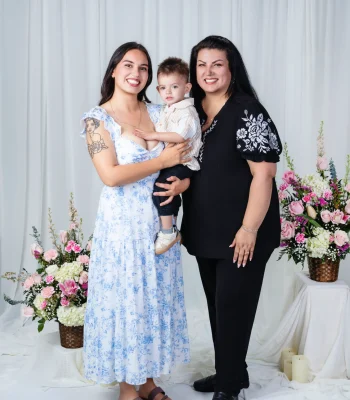 Young mother in a blue floral dress holding her toddler son beside the grandmother in a black top with white floral embroidery, in a white studio with pink flowers.
