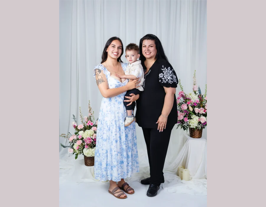 Young mother in a blue floral dress holding her toddler son beside the grandmother in a black top with white floral embroidery, in a white studio with pink flowers.