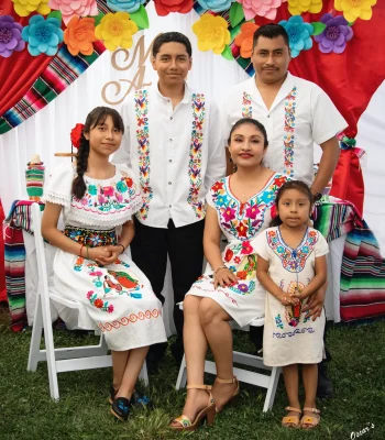 Family of five in traditional Mexican embroidered clothing, posed at an outdoor celebration with colorful paper flowers and striped sarape fabric behind them.
