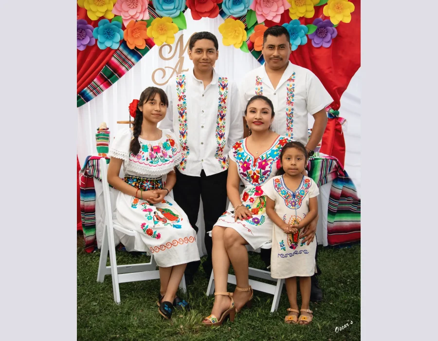 Family of five in traditional Mexican embroidered clothing, posed at an outdoor celebration with colorful paper flowers and striped sarape fabric behind them.