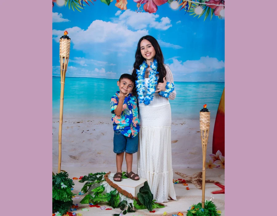 Young woman in a white lace dress and blue flower lei with a little boy in a Hawaiian shirt, both making shaka signs on a tropical beach set with tiki torches at Oscar's Photography and Video.