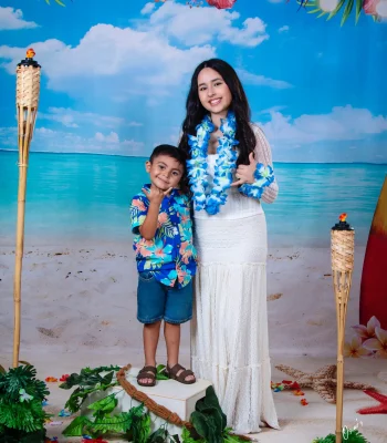 Young woman in a white lace dress and blue flower lei with a little boy in a Hawaiian shirt, both making shaka signs on a tropical beach set with tiki torches at Oscar's Photography and Video.