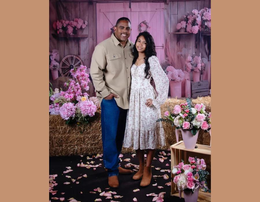 Father in a khaki shirt and jeans standing with his teen daughter in a floral midi dress, on a rustic pink barn set with hay bales and pink flowers.