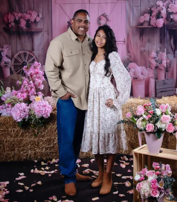 Father in a khaki shirt and jeans standing with his teen daughter in a floral midi dress, on a rustic pink barn set with hay bales and pink flowers.