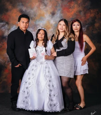 Girl in a white first communion gown and floral tiara holding a prayer book and candle, posed with her father and two older sisters against a painted backdrop.