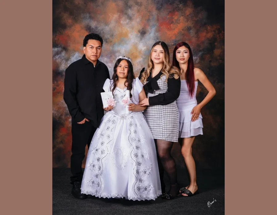 Girl in a white first communion gown and floral tiara holding a prayer book and candle, posed with her father and two older sisters against a painted backdrop.