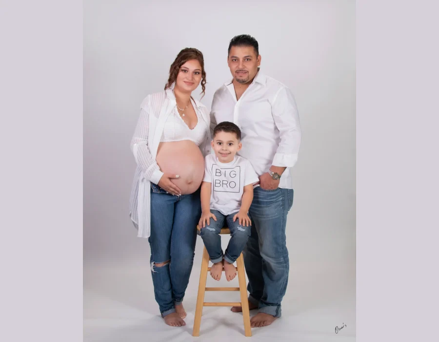 Maternity family portrait of an expecting mother in white and jeans, a father in a white shirt, and their son in a "Big Bro" tee seated on a wooden stool.