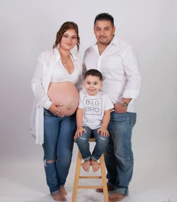 Maternity family portrait of an expecting mother in white and jeans, a father in a white shirt, and their son in a "Big Bro" tee seated on a wooden stool.