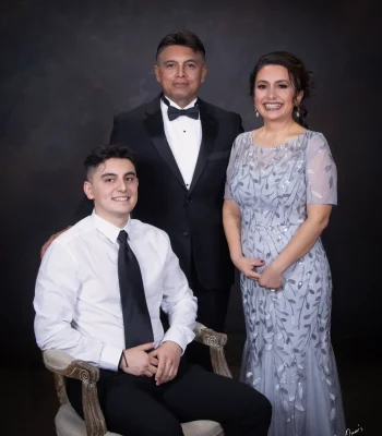 Family portrait of a smiling young man in a white shirt and black tie seated in an armchair, with his father in a tuxedo and mother in a silver beaded gown standing behind him.