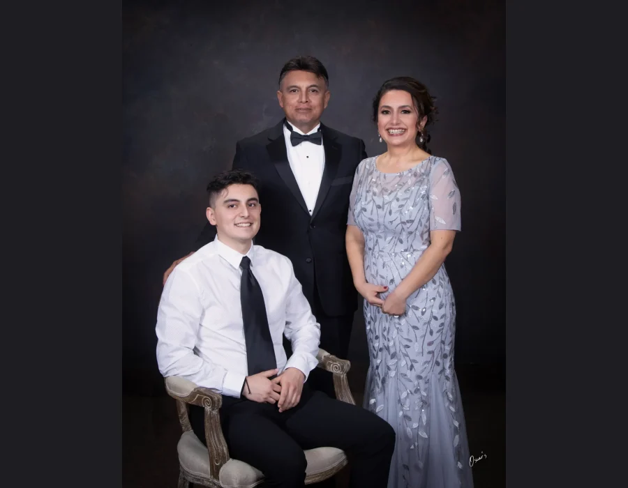 Family portrait of a smiling young man in a white shirt and black tie seated in an armchair, with his father in a tuxedo and mother in a silver beaded gown standing behind him.