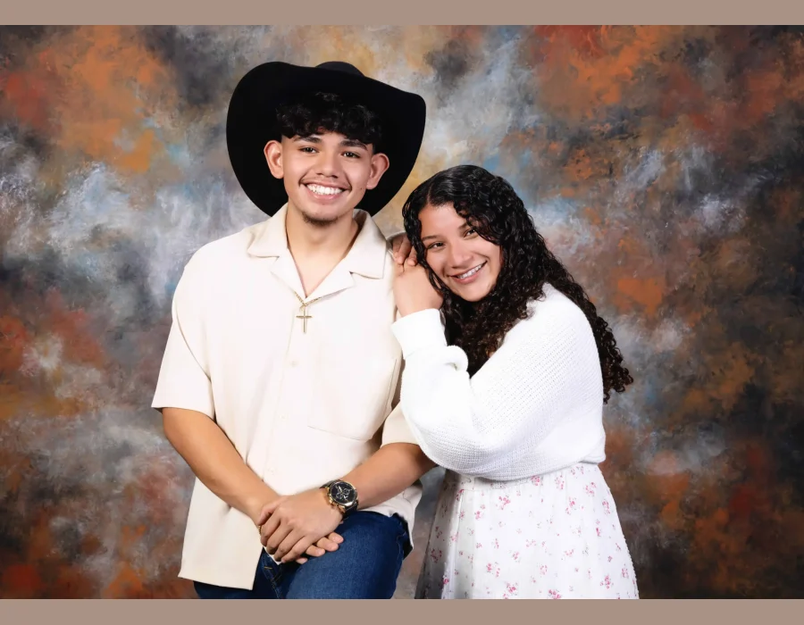 Smiling young man in a black cowboy hat, cream button-up shirt and gold cross necklace, with a curly-haired young woman in a white knit cardigan resting her head on his shoulder, against a painted backdrop.