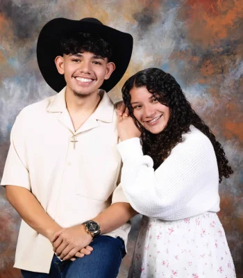 Smiling young man in a black cowboy hat, cream button-up shirt and gold cross necklace, with a curly-haired young woman in a white knit cardigan resting her head on his shoulder, against a painted backdrop.