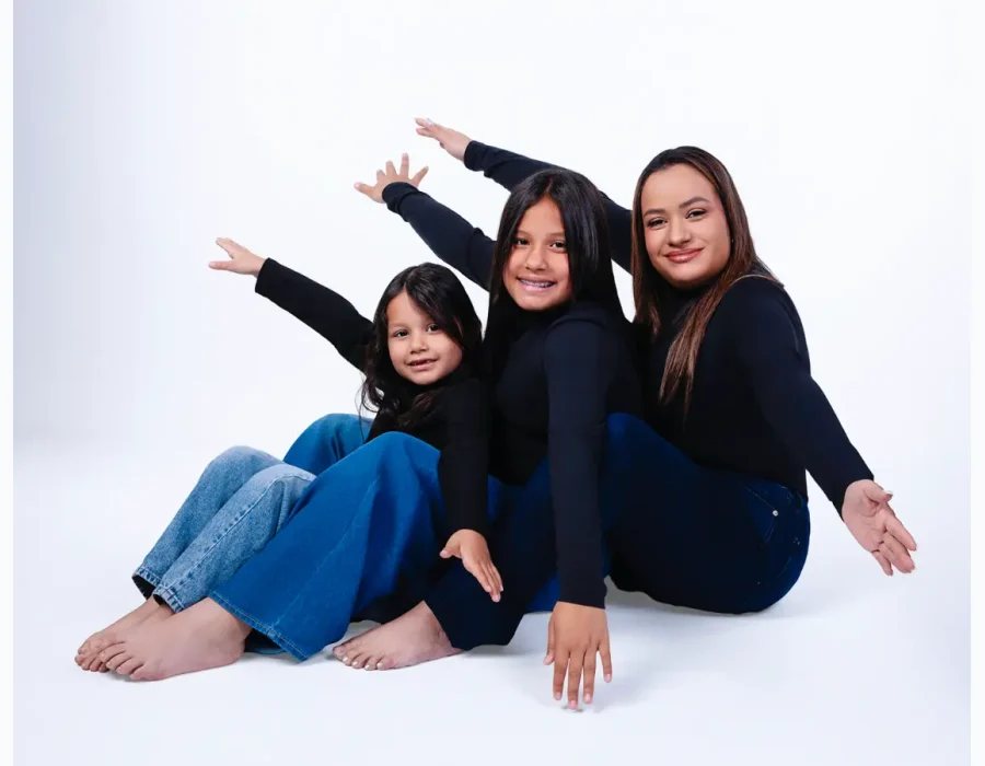 Mother and two daughters in matching black turtlenecks and jeans, sitting barefoot on a white studio floor with their arms stretched out in a playful staggered pose.