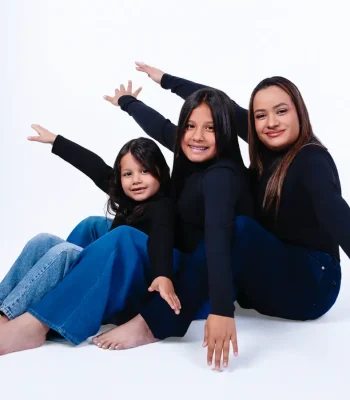 Mother and two daughters in matching black turtlenecks and jeans, sitting barefoot on a white studio floor with their arms stretched out in a playful staggered pose.