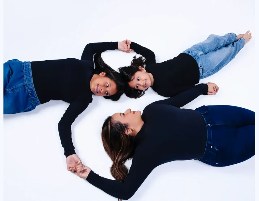 Overhead view of a mother and two daughters in black tops and jeans lying on a white floor, heads close together and holding hands in a circle, at Oscar's Photography and Video.