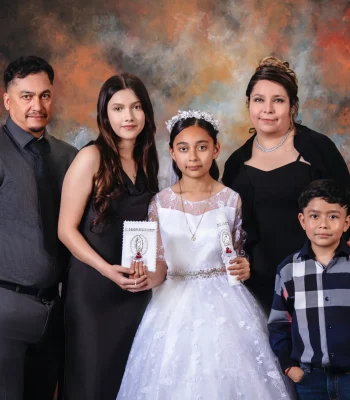 First communion family portrait of a girl in a white lace gown and floral crown holding a candle, with her father, older sister holding a prayer book, mother, and younger brother in a plaid shirt.