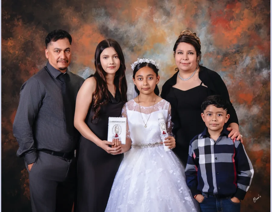 First communion family portrait of a girl in a white lace gown and floral crown holding a candle, with her father, older sister holding a prayer book, mother, and younger brother in a plaid shirt.
