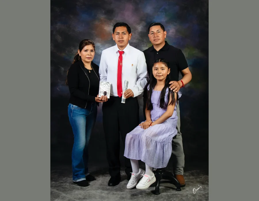 Religious celebration portrait of a teen son in a white shirt and red tie holding a ceremonial candle, with his mother holding a silver prayer book, his father, and his sister in a lavender dress seated on a stool.