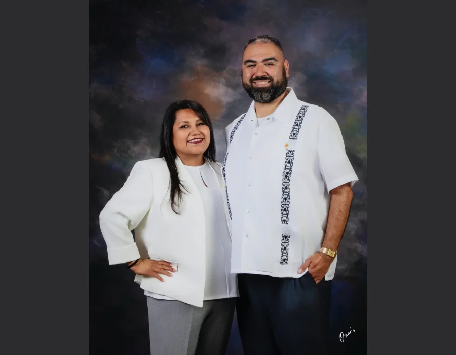 Studio portrait of a smiling couple, the woman in a white blazer and gray trousers with a hand on her hip, and the bearded man in a white embroidered guayabera, against a dark painted backdrop.