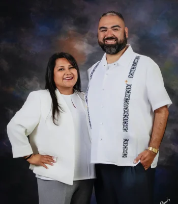 Studio portrait of a smiling couple, the woman in a white blazer and gray trousers with a hand on her hip, and the bearded man in a white embroidered guayabera, against a dark painted backdrop.