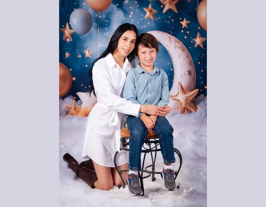 Mother in a white shirt dress kneeling behind her son seated on a stool, posed on a whimsical set with a crescent moon, gold stars, balloons, and clouds.