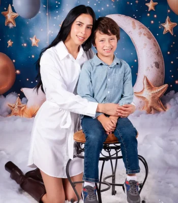 Mother in a white shirt dress kneeling behind her son seated on a stool, posed on a whimsical set with a crescent moon, gold stars, balloons, and clouds.