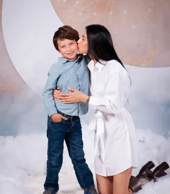 Mother in a white shirt dress kneeling and kissing her smiling son's cheek on a dreamy cloud set with a large pastel moon backdrop at Oscar's Photography and Video.