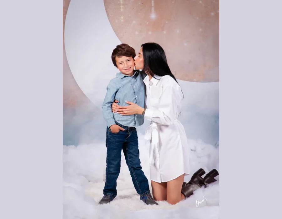 Mother in a white shirt dress kneeling and kissing her smiling son's cheek on a dreamy cloud set with a large pastel moon backdrop at Oscar's Photography and Video.