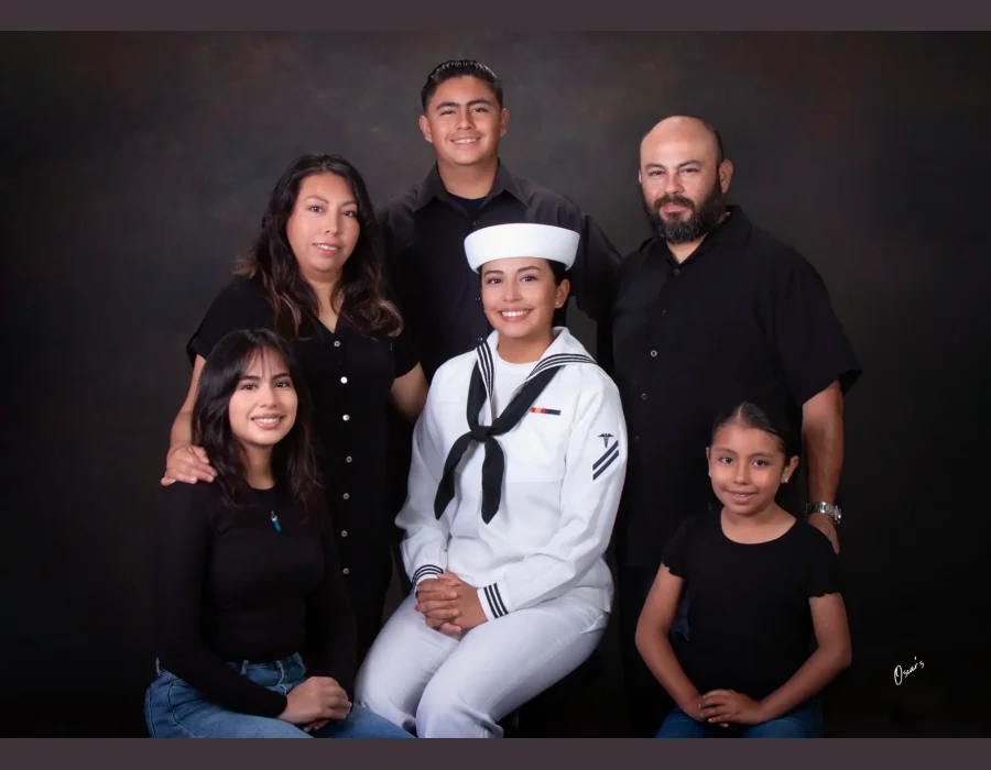 Family portrait celebrating a daughter in a U.S. Navy white dress uniform, seated at center and surrounded by her parents and three siblings dressed in black.