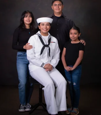 Young woman in a U.S. Navy white dress uniform seated on a stool, surrounded by her sister, brother, and younger sister in black tops and jeans.