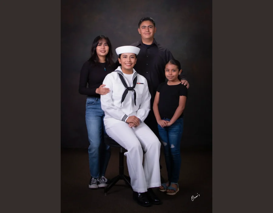 Young woman in a U.S. Navy white dress uniform seated on a stool, surrounded by her sister, brother, and younger sister in black tops and jeans.