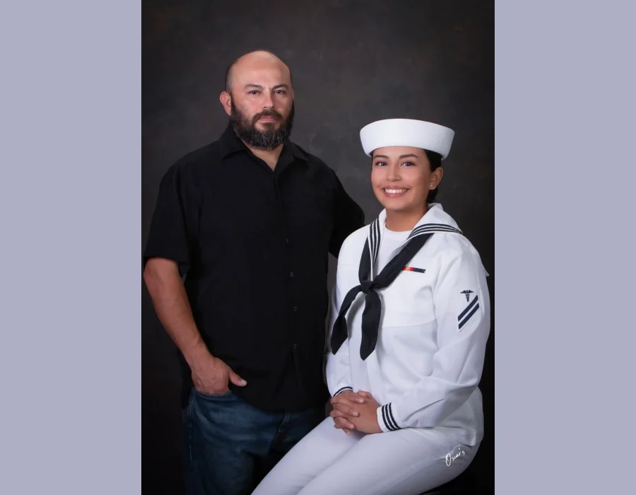 Father in a black shirt standing beside his smiling daughter, who is seated in a U.S. Navy white dress uniform and cap, against a dark textured backdrop.