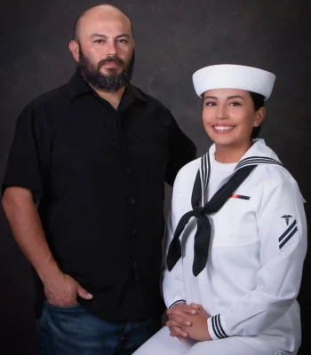 Father in a black shirt standing beside his smiling daughter, who is seated in a U.S. Navy white dress uniform and cap, against a dark textured backdrop.