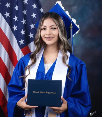 Graduation portrait of a girl in blue cap and gown holding her diploma in front of an American flag, studio backdrop