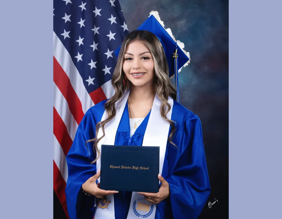Graduation portrait of a girl in blue cap and gown holding her diploma in front of an American flag, studio backdrop