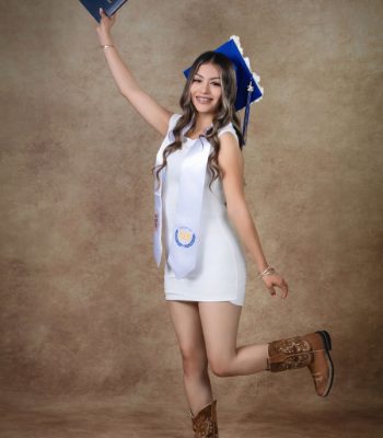 Graduation portrait of a girl in a white dress and cowboy boots jumping with her diploma raised, Class of 2026 sash