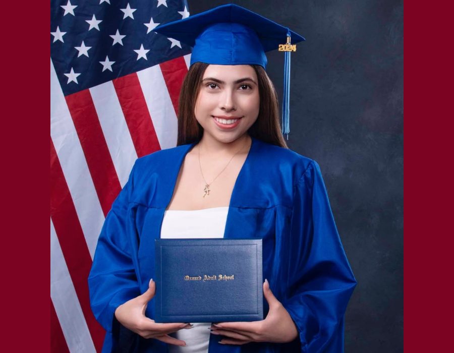 Graduation portrait of a young woman in blue cap and gown holding her diploma in front of an American flag, studio backdrop