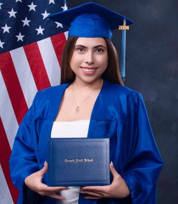 Graduation portrait of a young woman in blue cap and gown holding her diploma in front of an American flag, studio backdrop