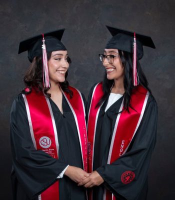 Two college graduates in cap and gown with red honor stoles holding hands, studio graduation portrait