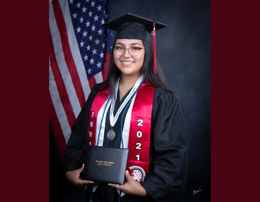Smiling high school graduate in glasses, a black cap and gown, and a red "RMHS 2021" stole with a medal, holding a Rio Mesa High School diploma in front of an American flag.