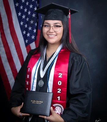Smiling high school graduate in glasses, a black cap and gown, and a red "RMHS 2021" stole with a medal, holding a Rio Mesa High School diploma in front of an American flag.