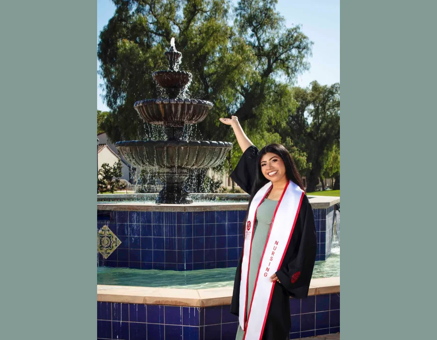 Nursing graduate in a black gown and white "Nursing" stole, smiling and gesturing toward a tiered fountain on a sunny campus.