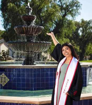 Nursing graduate in a black gown and white "Nursing" stole, smiling and gesturing toward a tiered fountain on a sunny campus.
