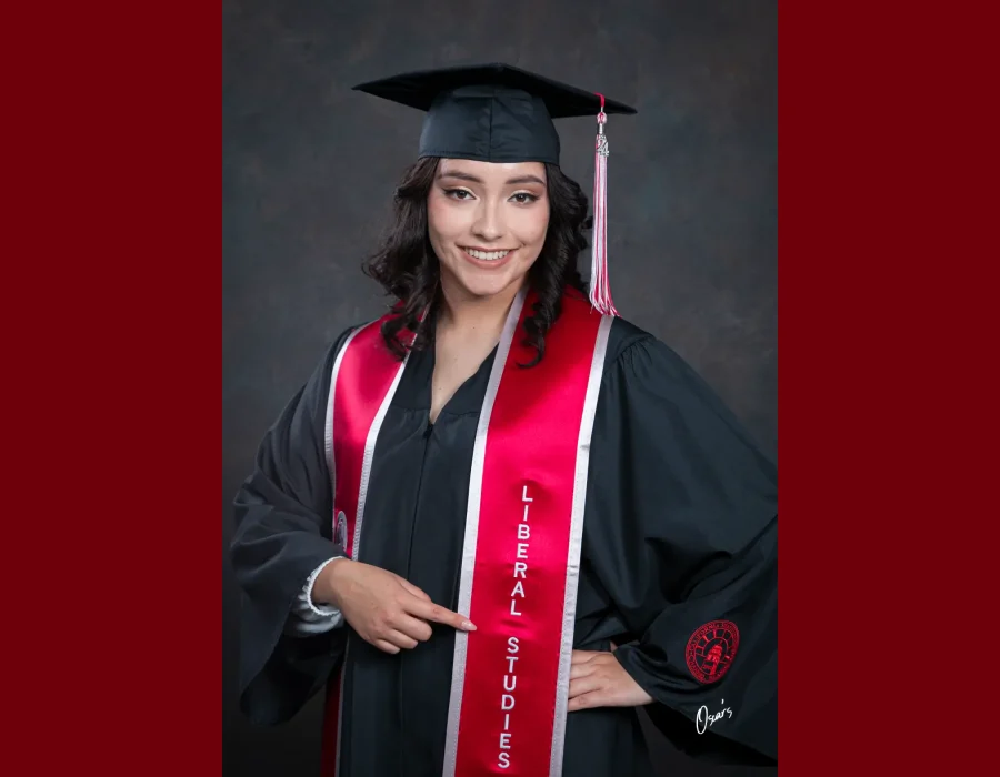 Graduate with curly dark hair in a black cap and gown and a red "Liberal Studies" stole, pointing to her major with a hand on her hip.