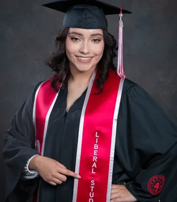 Graduate with curly dark hair in a black cap and gown and a red "Liberal Studies" stole, pointing to her major with a hand on her hip.