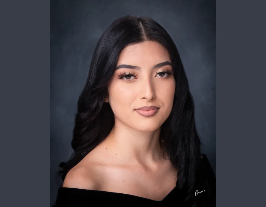 Classic senior portrait of a young woman with long black hair and soft glam makeup, wearing an off-the-shoulder black drape against a dark backdrop.