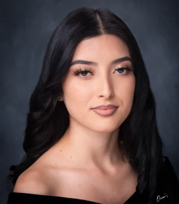 Classic senior portrait of a young woman with long black hair and soft glam makeup, wearing an off-the-shoulder black drape against a dark backdrop.
