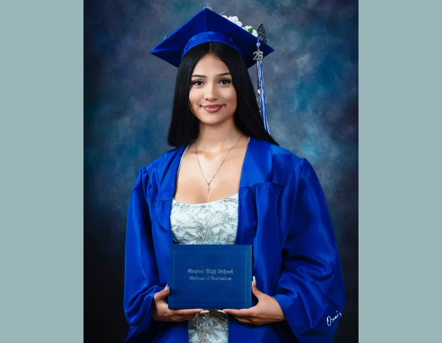 Graduate with long black hair in a blue gown and a flower-decorated cap with a "26" tassel charm, holding a Condor High School diploma.
