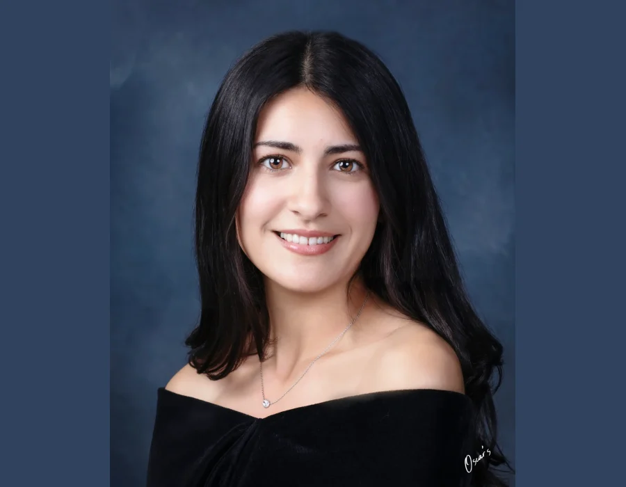 Senior portrait of a smiling young woman with long dark hair and a delicate pendant necklace, wearing an off-the-shoulder black drape against a blue backdrop.